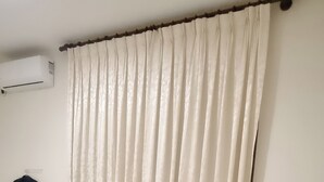 En pænt redt seng med hvide sengetæppe og puder, en mørk jakke hængt på en krog, og en hvid aircondition-enhed på væggen.