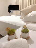 Una cama bien tendida con ropa de cama blanca, dos pequeñas macetas con plantas sobre una mesita de noche y una toalla negra colgada en un perchero.