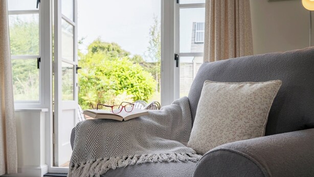 A grey sofa with a patterned pillow and a knitted throw, a pair of glasses on an open book, and a view of greenery through a glass door. - Hollybank Cottage, Ufford (Ufford)