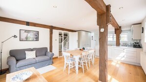 A spacious living area with a wooden dining table, white chairs, a grey sofa, and a kitchen in the background. - Hollybank Cottage, Ufford (Ufford)