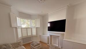 Un salon doté d’un grand téléviseur à écran plat fixé au-dessus d’une cheminée, d’un canapé d’angle et d’une fenêtre avec des volets.
