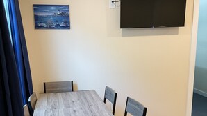 Et spiseområde med et træbord og stole, et fladskærms-tv ophængt på væggen og et maleri af et maritimt landskab.