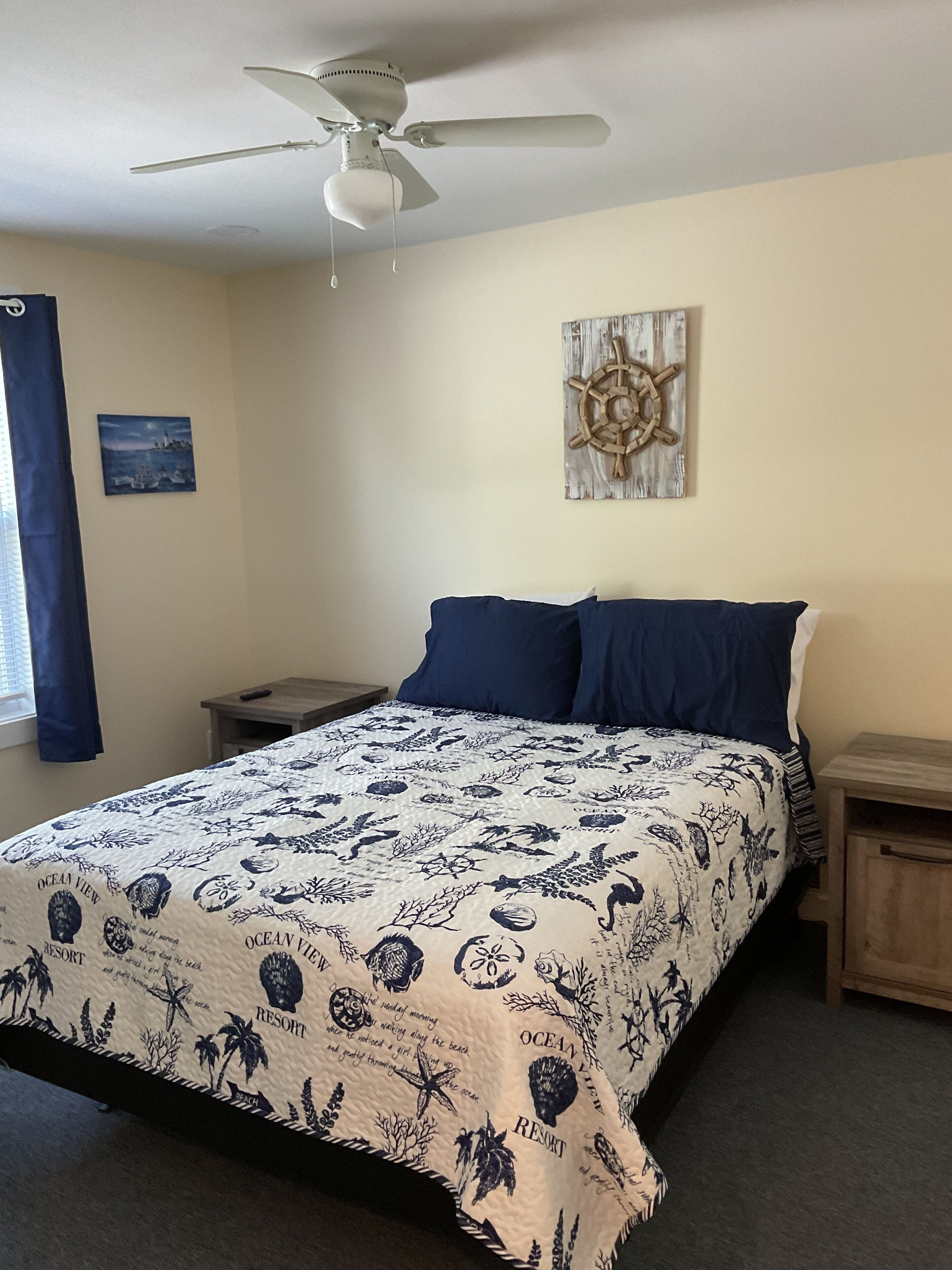 A bedroom with a bed, a ceiling fan, a nightstand, a wall clock, and a painting.
