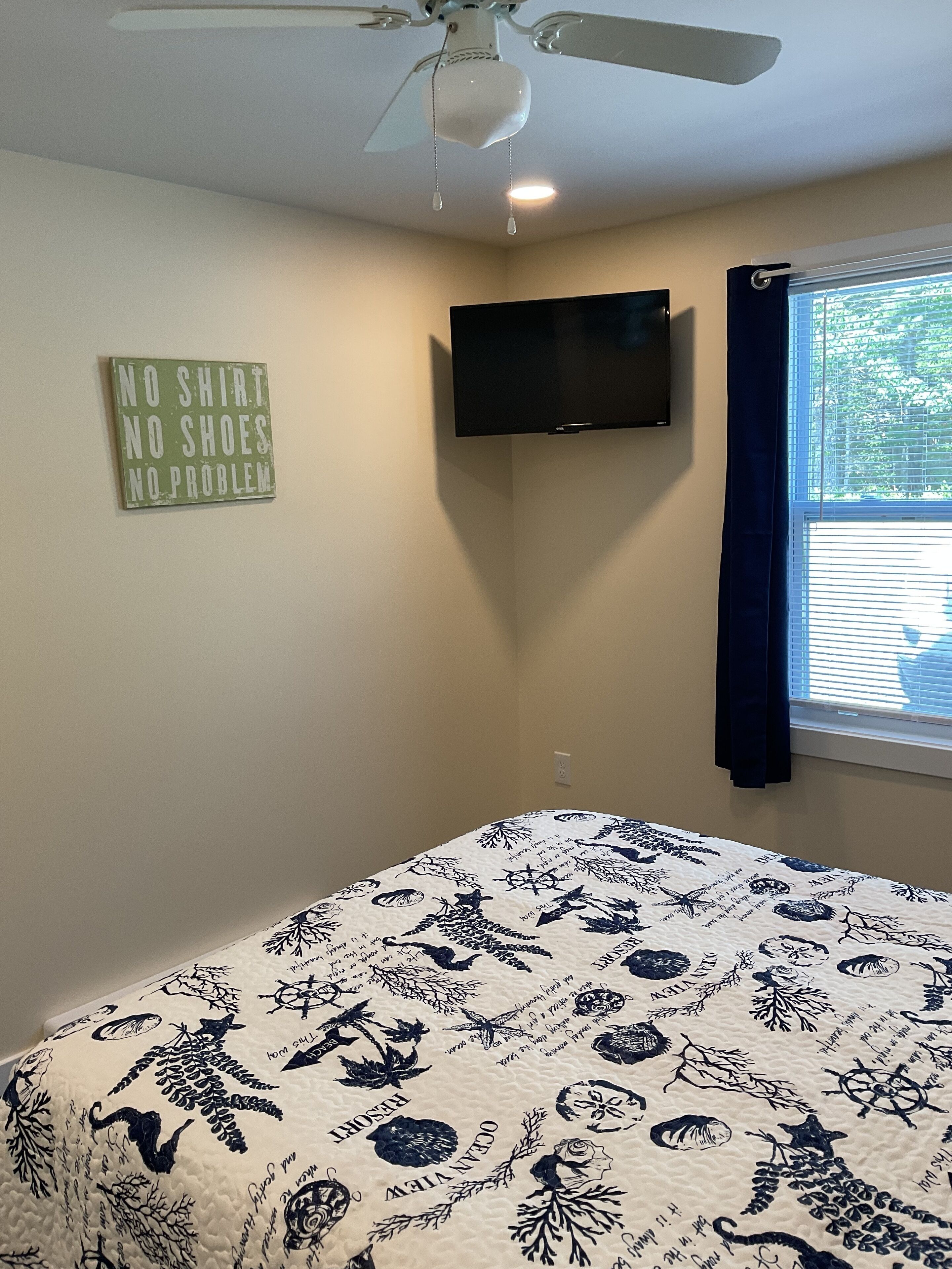 Een slaapkamer met een bed, een plafondventilator, een aan de muur bevestigde televisie en een raam met gordijnen.