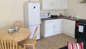 A compact kitchen with white cabinets, a microwave, a kettle, and a stove. There is a wooden dining table with chairs and a small black couch with a striped pillow.