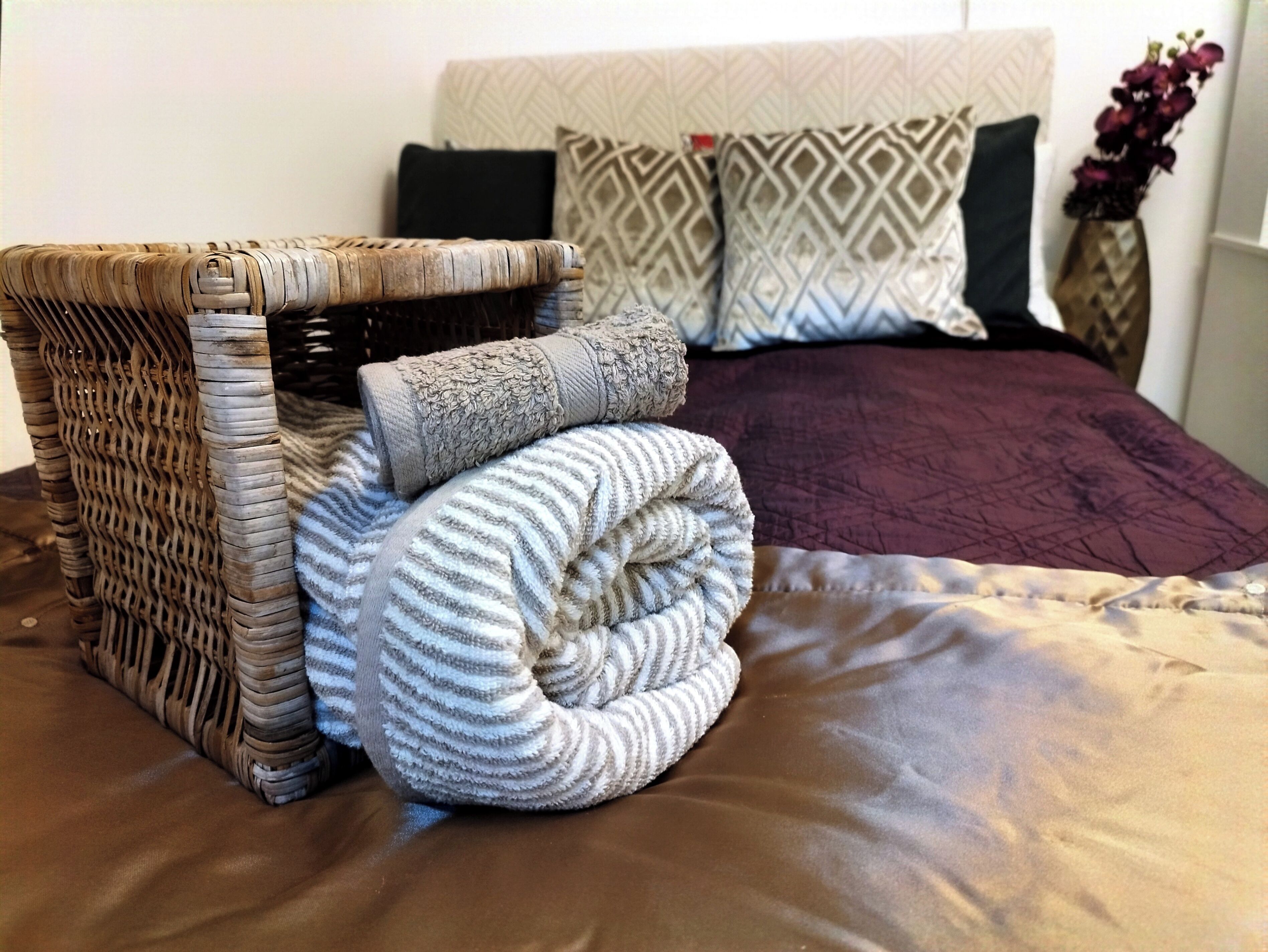 Ein Bett mit einem gemusterten Kopfteil, ein gewebter Nachttisch und eine zusammenrollte Decke.