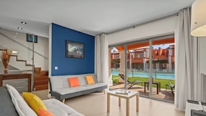 A modern living room with a grey sofa, a blue accent wall, and a view of a swimming pool and buildings.