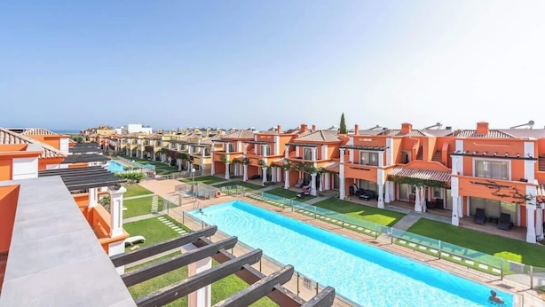A resort with multiple orange-roofed buildings, a central swimming pool, and a clear view of the ocean.
