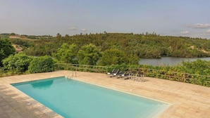 A rectangular swimming pool with a clear blue water surface, surrounded by a paved deck. There are lounge chairs and a small table on the deck, and a view of a forested area and a body of water in the background. - Quiet Escape by the River w/ Pool by HostWise (Tábua)