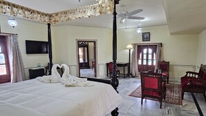 Une chambre d’hôtel avec un grand lit, un coin salon avec un fauteuil rouge, un ventilateur de plafond et un miroir qui reflète la pièce.