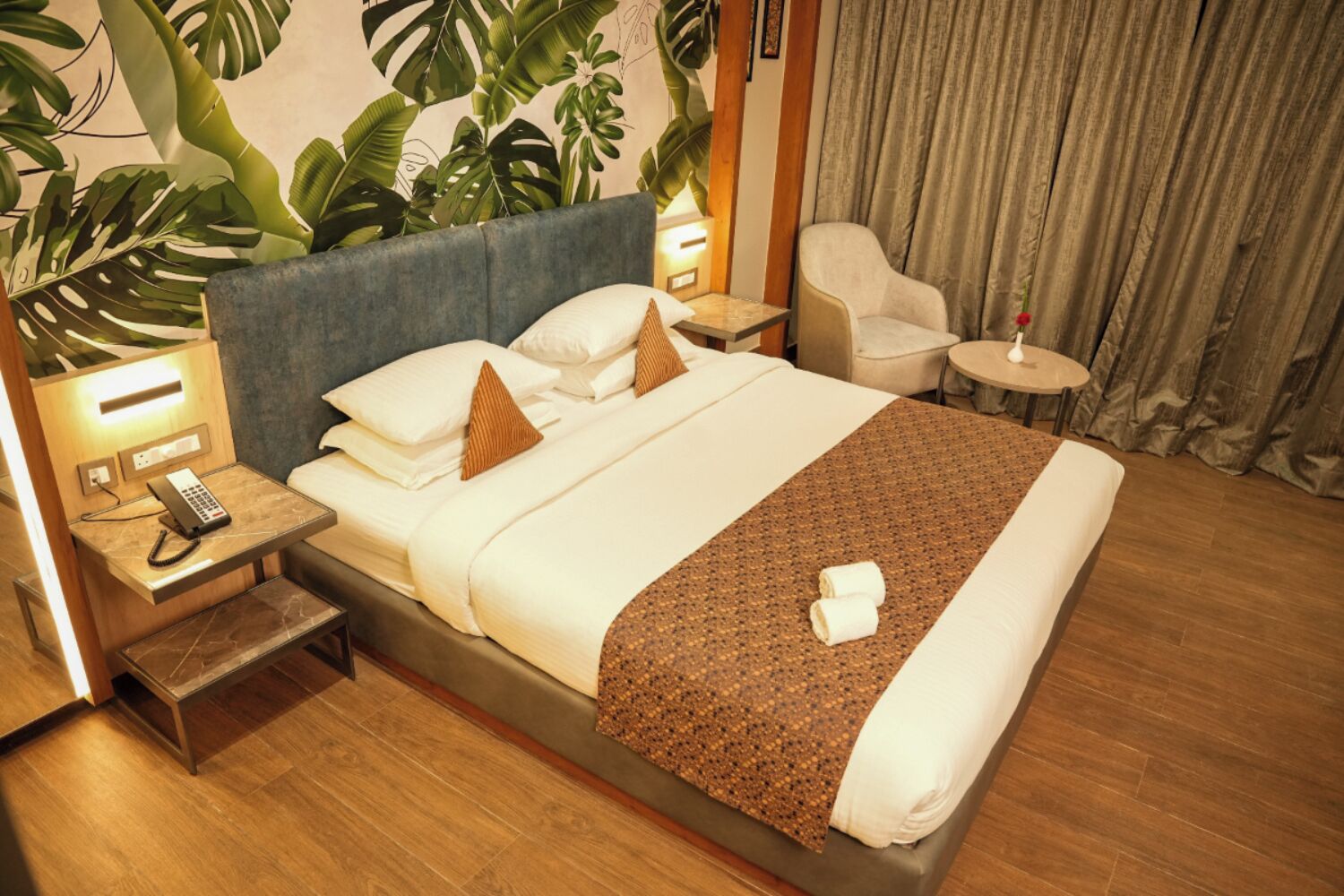 A hotel room with a large bed, a bedside table with a phone, a chair, a small table with a lamp, and a curtain.