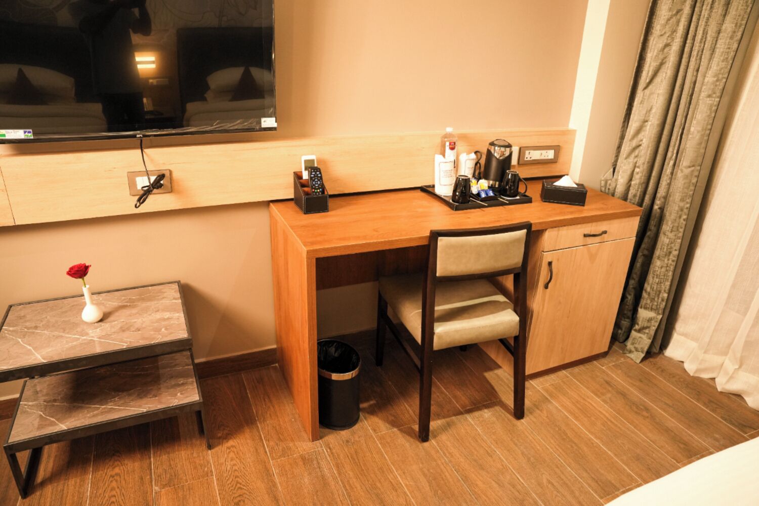 A compact hotel room with a wooden desk, a chair, a small table with a vase of a red rose, a trash bin, and a large window with a view of the room.
