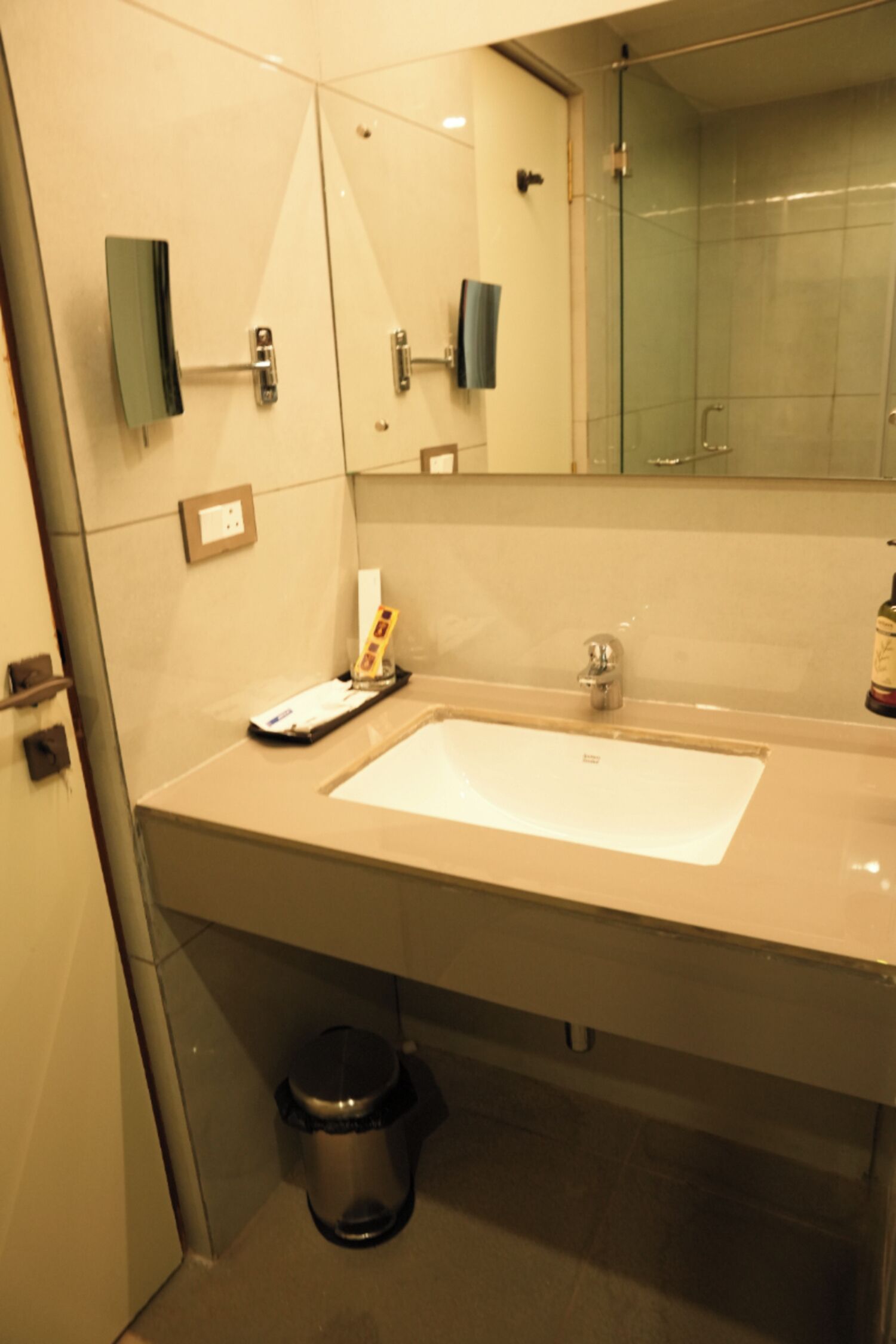 A bathroom with a large sink, a mirror, a glass shower enclosure, and a trash bin.