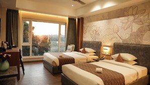 A hotel room with two beds, a desk, a chair, a large window with a view, and a ceiling fan. - Hotel Menaka Inn Dharwad (Dharwad)