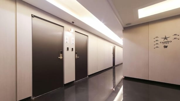 Un pasillo moderno de hotel con tres puertas de ascensor, suelo negro pulido y una pared con el nombre del hotel y el número de habitación.