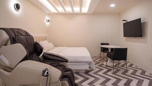 Habitación de hotel moderna con cama, sillón de masaje, una mesita y un televisor en la pared.