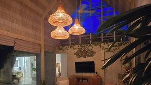 Un salon spacieux avec un grand canapé, une table basse et un téléviseur posé sur un meuble en bois. Trois lampes suspendues en osier sont accrochées au plafond, et une grande fenêtre offre une vue sur l’extérieur.