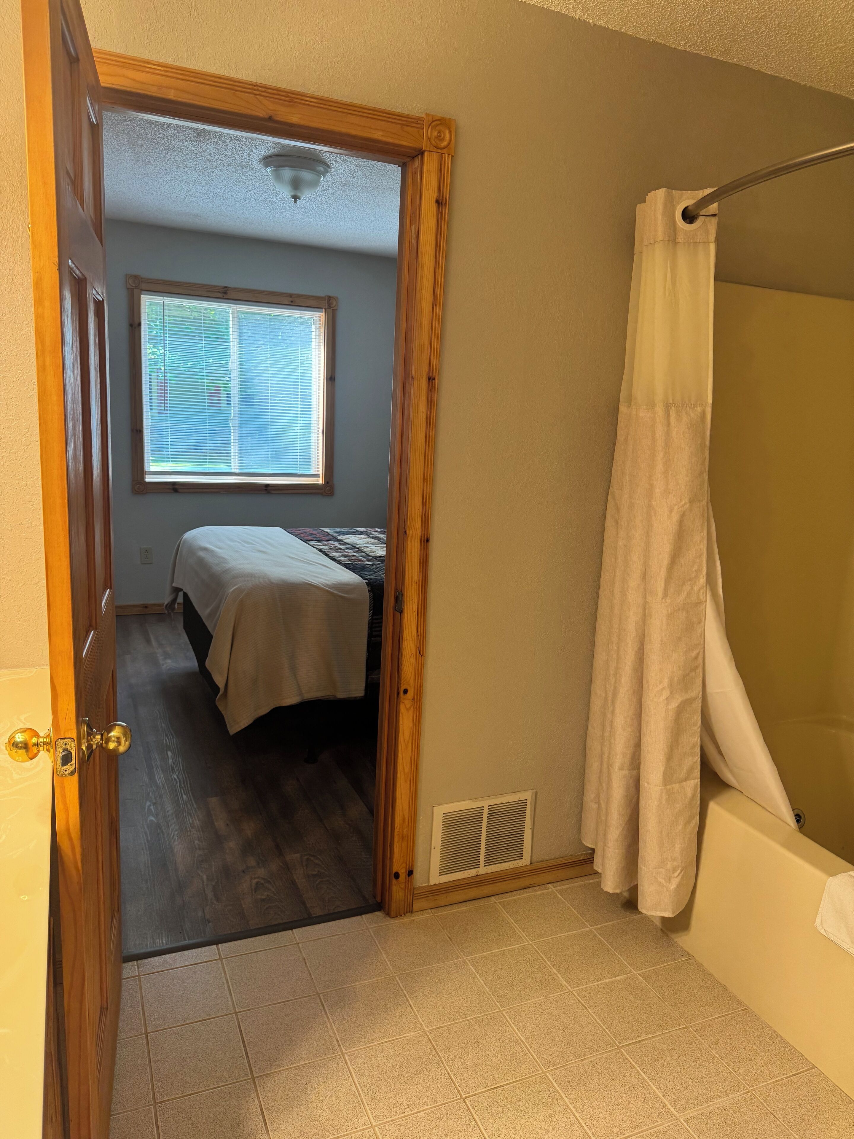 A bedroom with a bed, a window, a bathroom with a bathtub, and a door leading to another room.