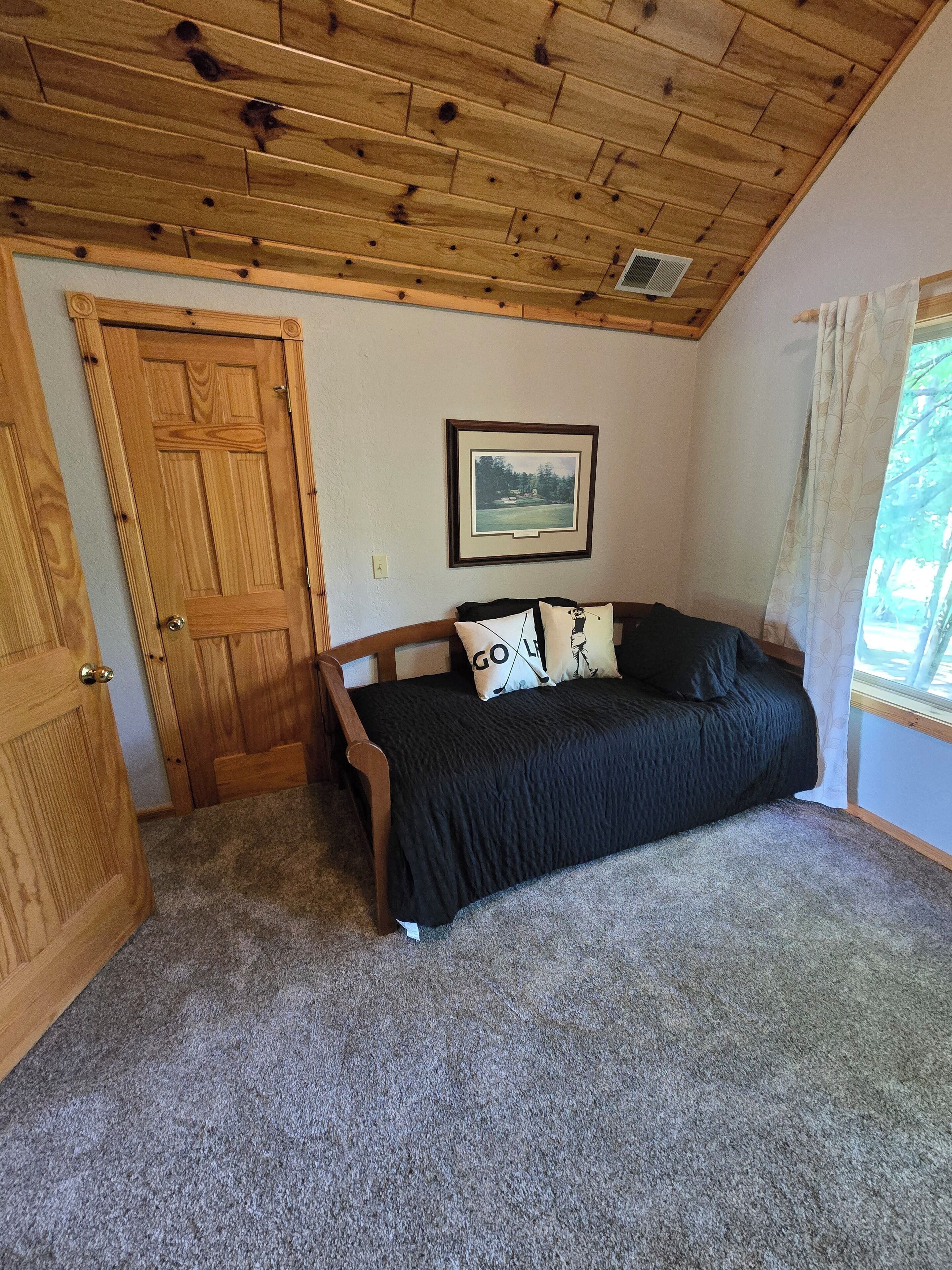 A room with a wooden ceiling, a door, a framed picture on the wall, and a bed with pillows.