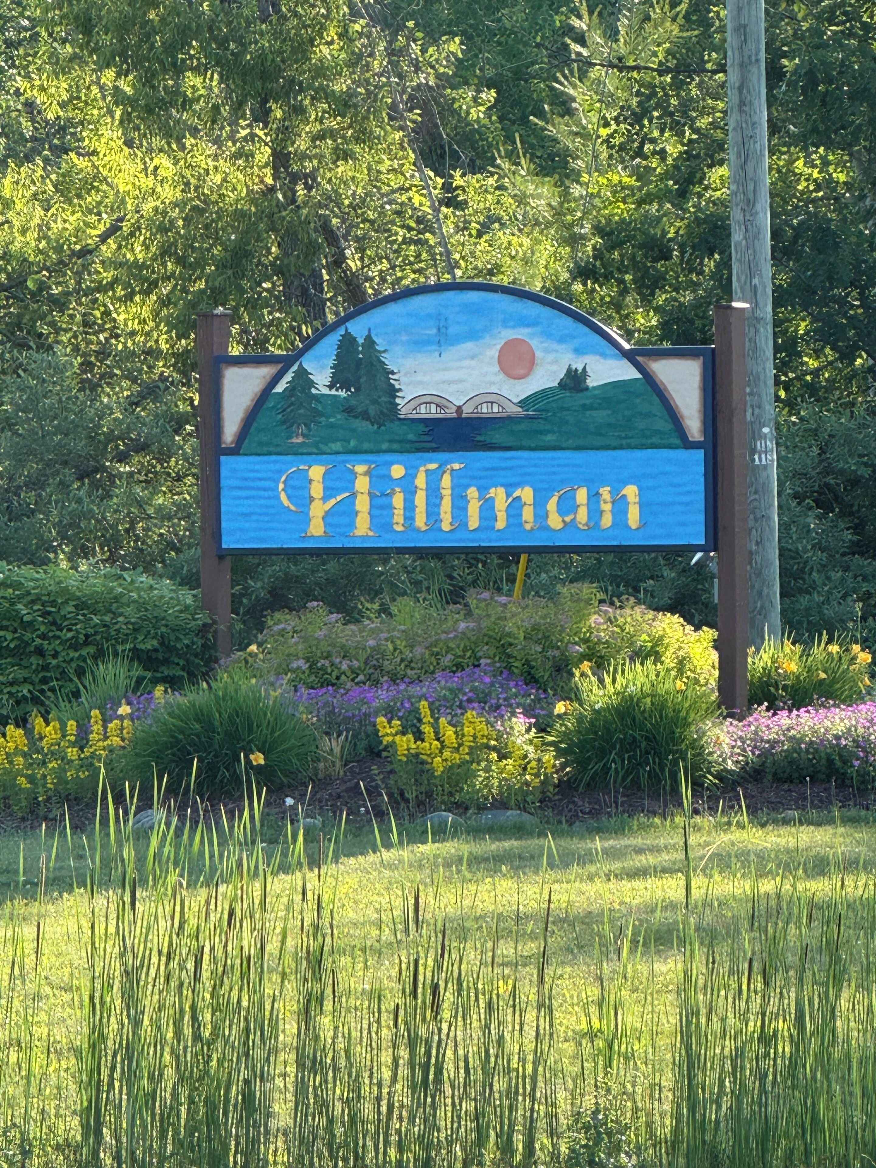 Une enseigne portant le nom « Hillman », entourée d’une végétation luxuriante et de fleurs.