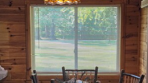 Interior de una cabaña de madera con una mesa de comedor y sillas, un ventanal con persianas y una lámpara decorativa.