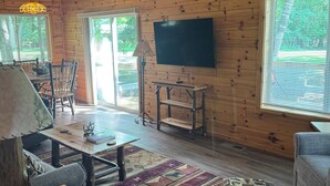 Interior acogedor de una cabaña con paredes de madera, un televisor grande, una zona de comedor y vistas a través de las ventanas.