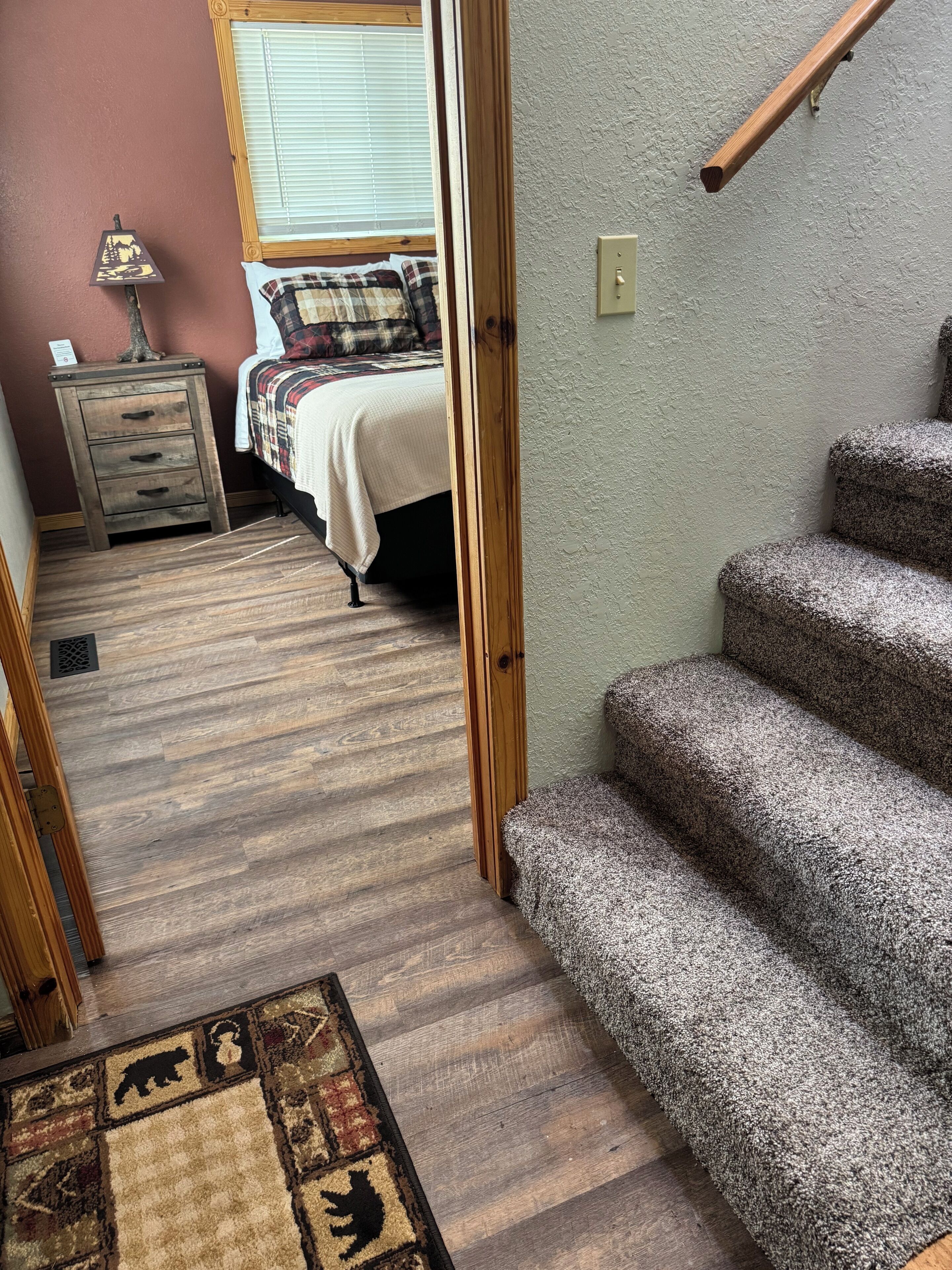 A hallway with a carpeted floor leading to a bedroom with a bed and a nightstand. There is a wooden staircase on the right.