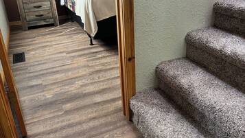 A hallway with a carpeted floor leading to a bedroom with a bed and a nightstand. There is a wooden staircase on the right.
