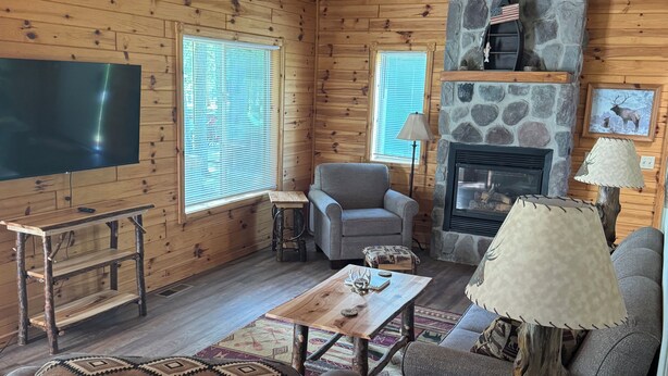 Acogedor interior de cabaña de madera con chimenea de piedra, televisor de pantalla plana y asientos cómodos.