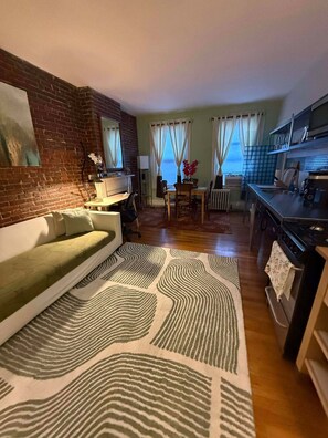 Ein Wohnzimmer mit einer Couch, einem Esstisch mit Stühlen, einer Küchenzeile mit Herd und Blick auf das Meer durch die Fenster.