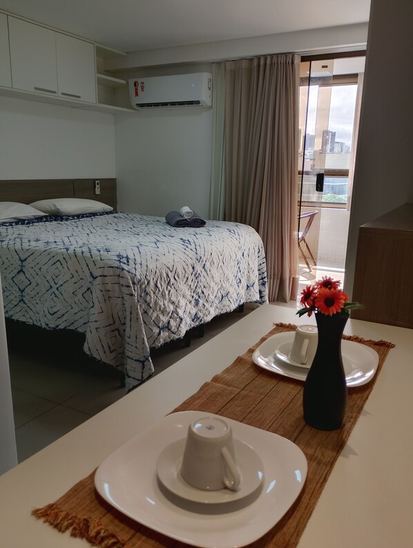 A hotel room with a bed, a table with a vase of flowers, and a view of the city through the window. - Nomar VII (Joao Pessoa)