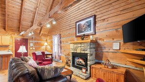 Interior acogedor de una cabaña de madera con chimenea de piedra, sofá de cuero, sillón rojo y televisor de pantalla plana montado en la pared.