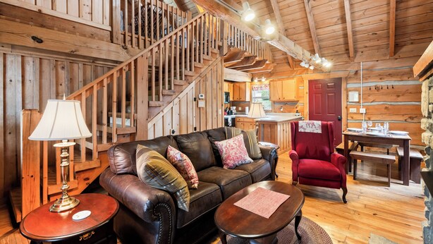 Interior acogedor de una cabaña de troncos con un sofá de cuero, un sillón rojo, una mesa de comedor y una escalera que conduce a un nivel superior.