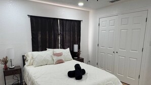 Ein Schlafzimmer mit einem Bett, einem Nachttisch, einer Lampe, einem schwarzen Stofftier, einem Fenster mit schwarzen Vorhängen und einer weißen Tür.