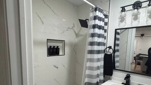 Ein Badezimmer mit weißer Marmorwand, einem schwarz-weißen Streifen-Duschvorhang, einem Waschbecken mit Spiegel darüber und einem Regal mit drei schwarzen Flaschen.