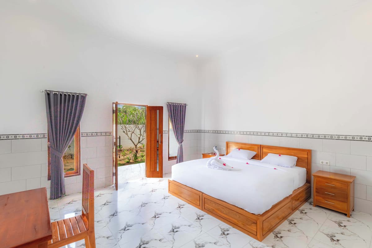 Un dormitorio con cama de madera, una mesita de noche, una silla y una pequeña mesa. Se ve una ventana con cortinas y vistas al exterior con vegetación.