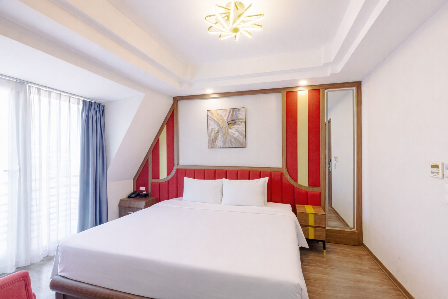 A hotel room with a large bed, a bedside table, a nightstand, a mirror, and a window with curtains.