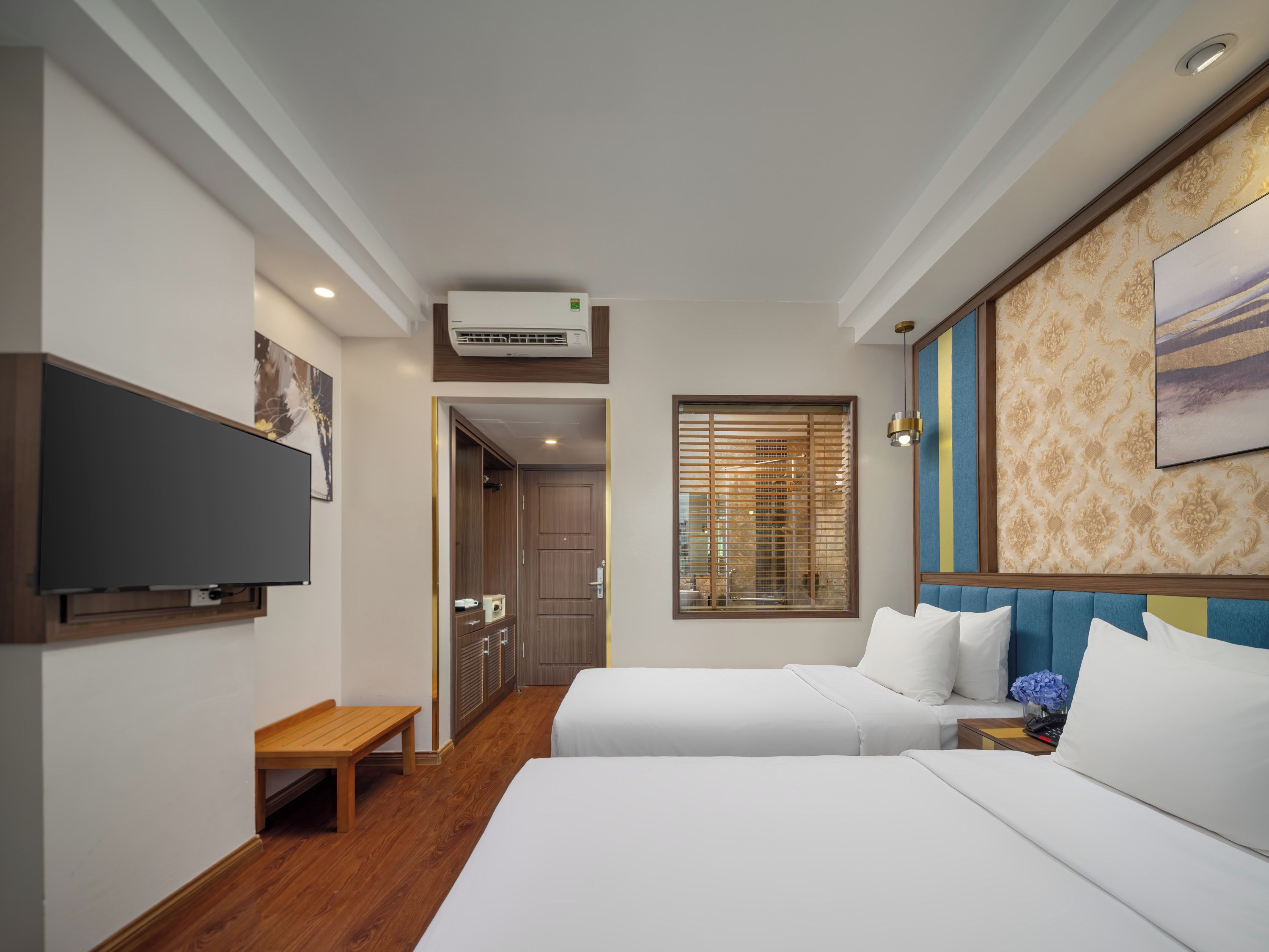 A hotel room with two beds, a flat-screen TV, a wooden bench, and a window with blinds.