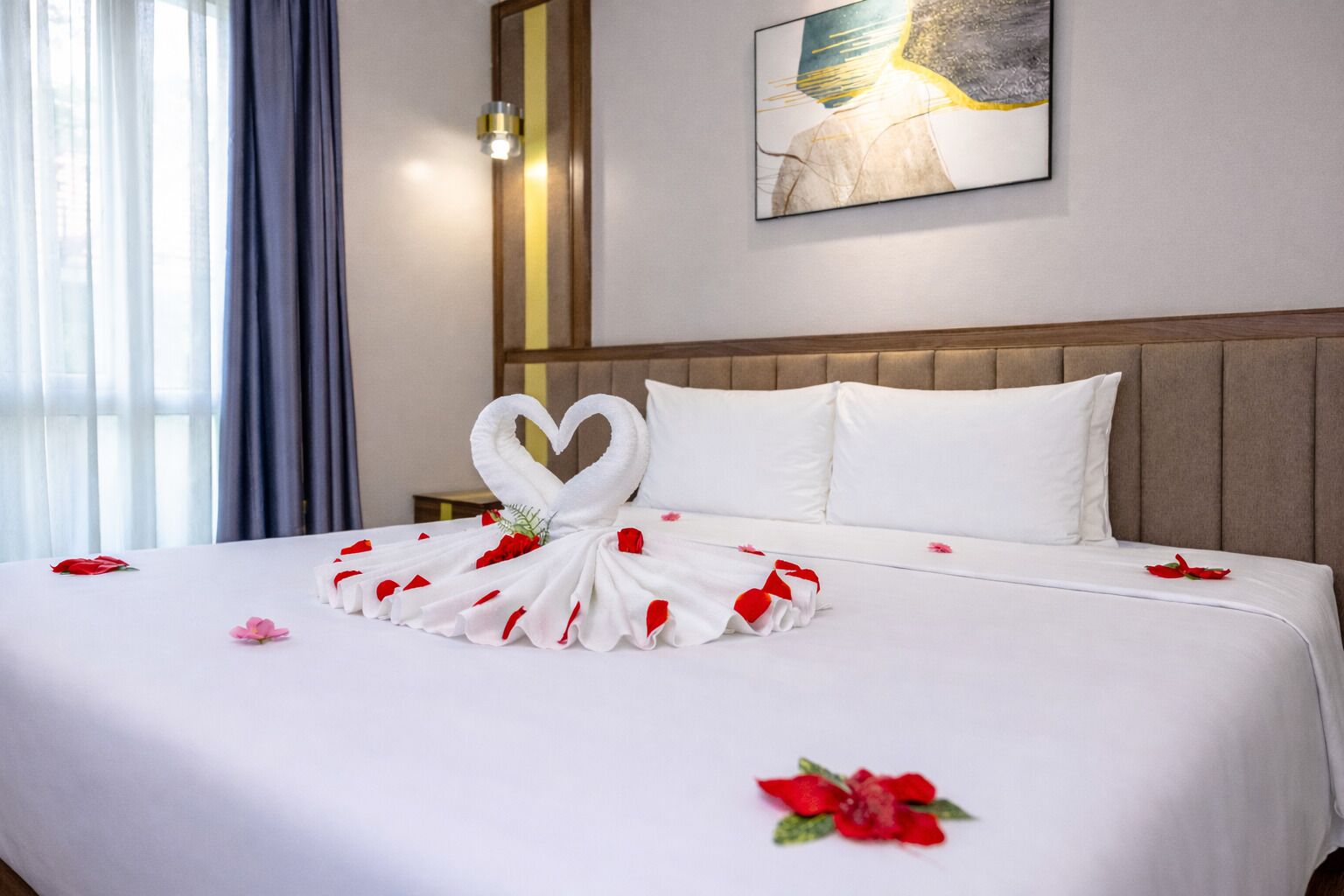 A hotel room with a neatly made bed, a heart-shaped towel arrangement, and a painting on the wall.