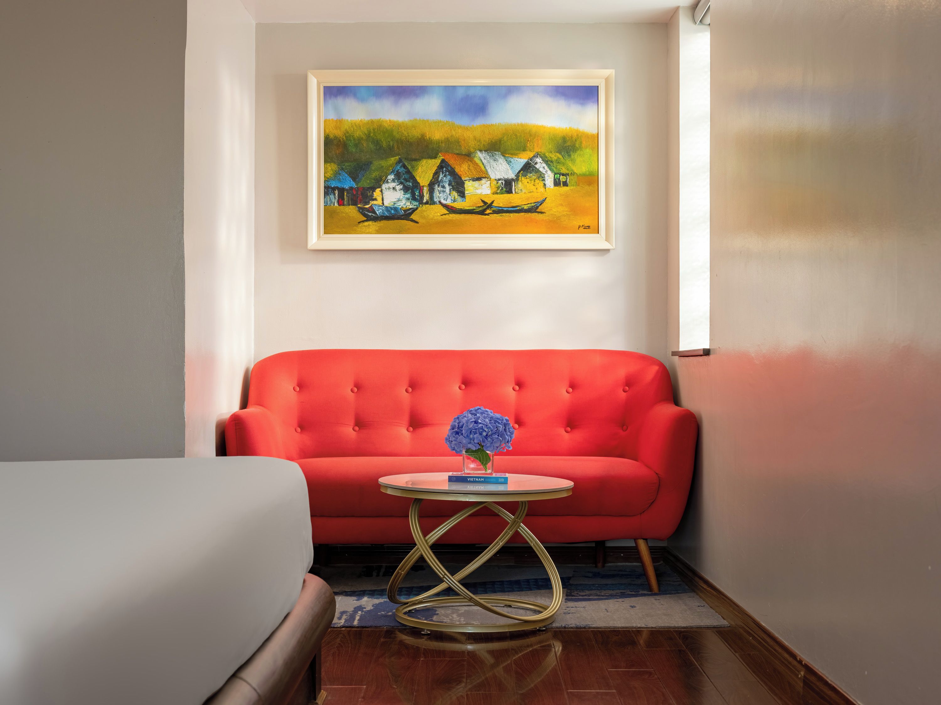 A room with a red sofa, a small round table with a vase of flowers, and a painting of houses on the wall.