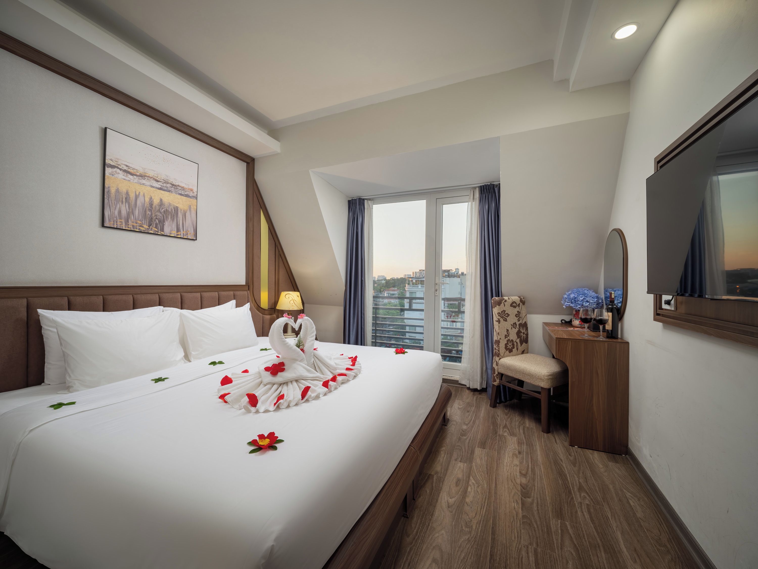 A hotel room with a large bed, a city view, a desk with a mirror, and a television.