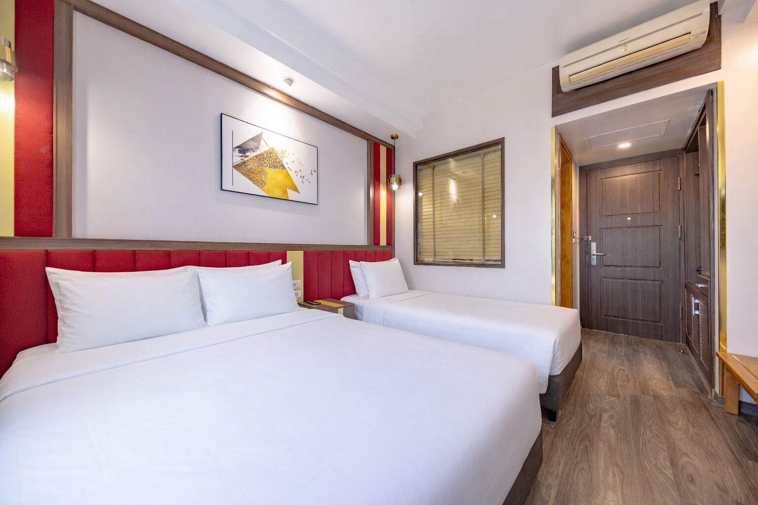 A hotel room with two beds, a red headboard, a wall-mounted artwork, a window with blinds, and an air conditioning unit.