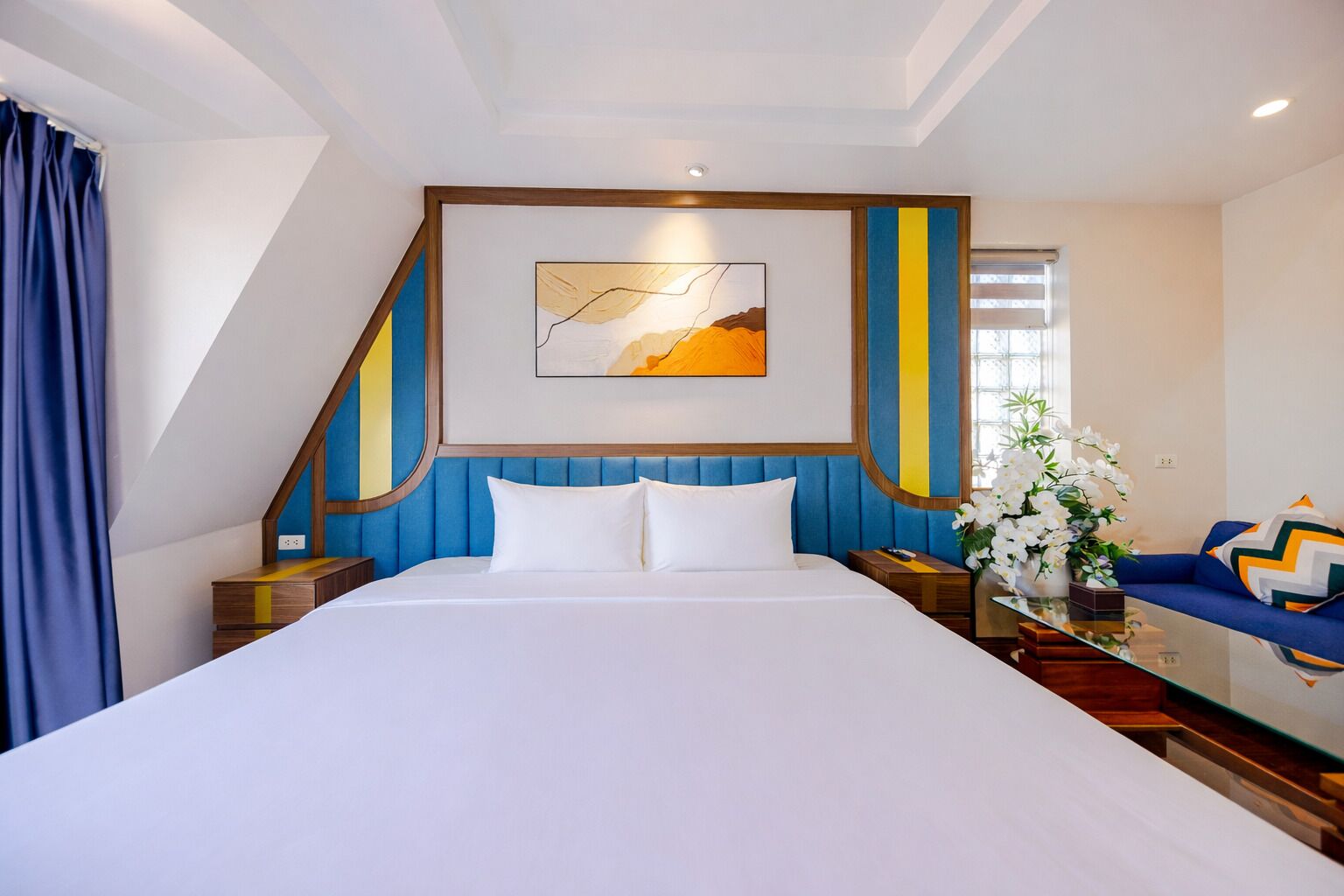 Habitación de hotel con una cama grande, cabecera azul, un cuadro sobre la cama, sofá con cojines estampados y un jarrón con flores.