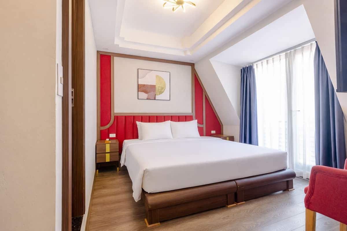 Une chambre d’hôtel avec un grand lit, une table de chevet en bois, un mur d’accent rouge et une fenêtre avec des rideaux.