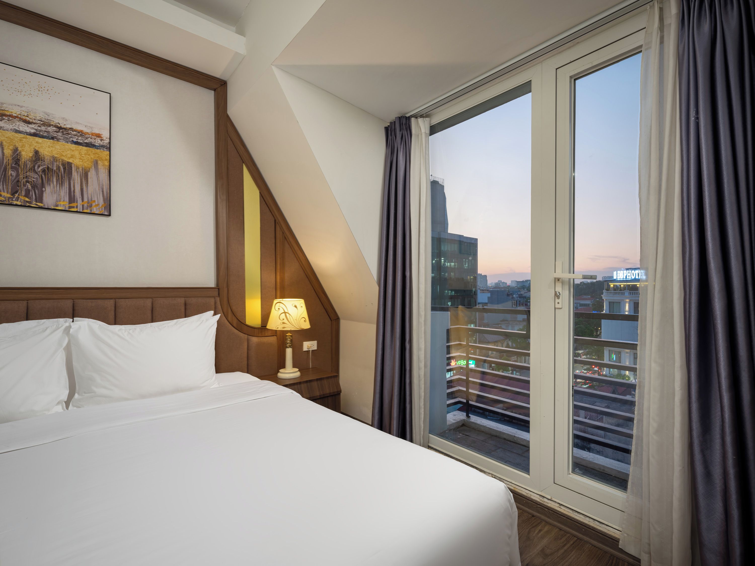 A hotel room with a bed, a nightstand, a lamp, and a large window with curtains.