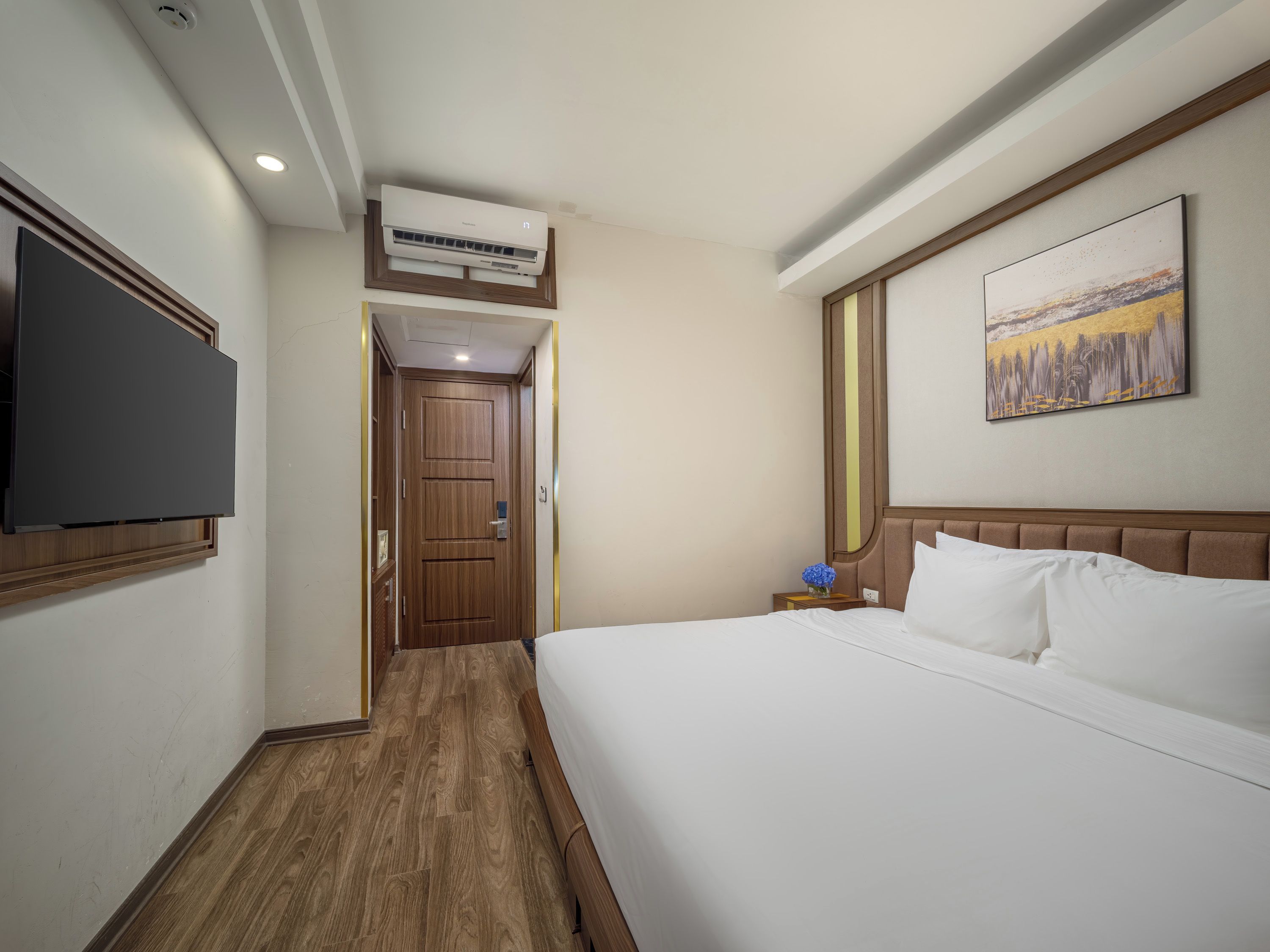 A hotel room with a bed, a flat-screen TV mounted on the wall, a wooden door, and a painting above the bed.