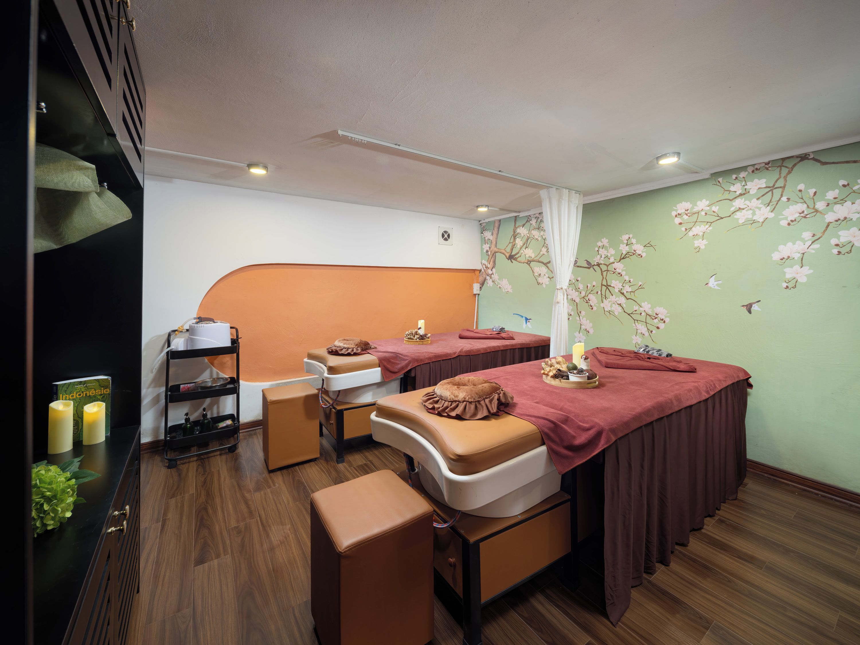 A room with two massage tables, a shelf with bottles, and a green wall with floral wallpaper.