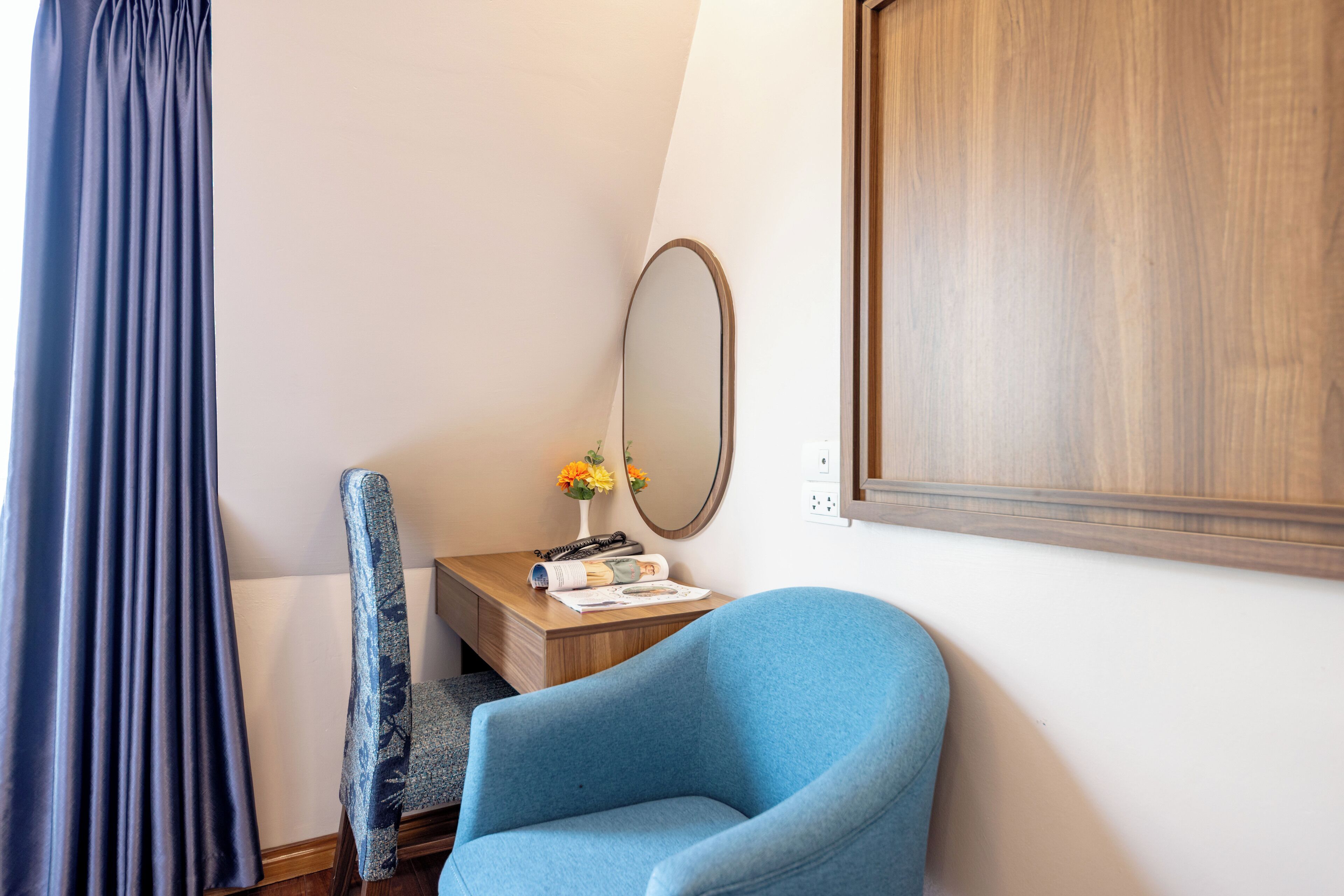 A room with a blue armchair, a wooden side table with a mirror, a vase with flowers, and a window with curtains.