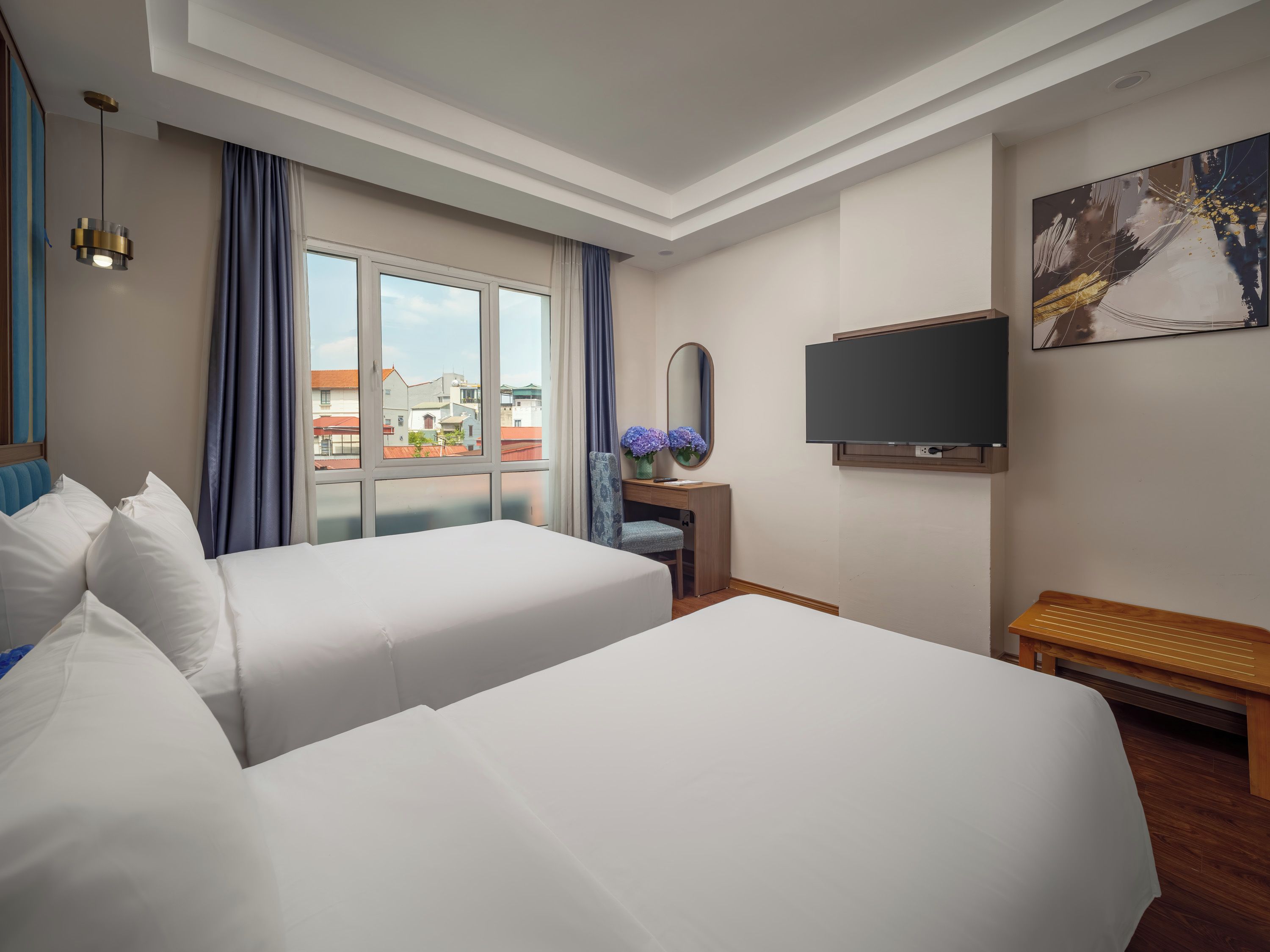 A hotel room with two beds, a desk, a TV, and a large window with a view of buildings.