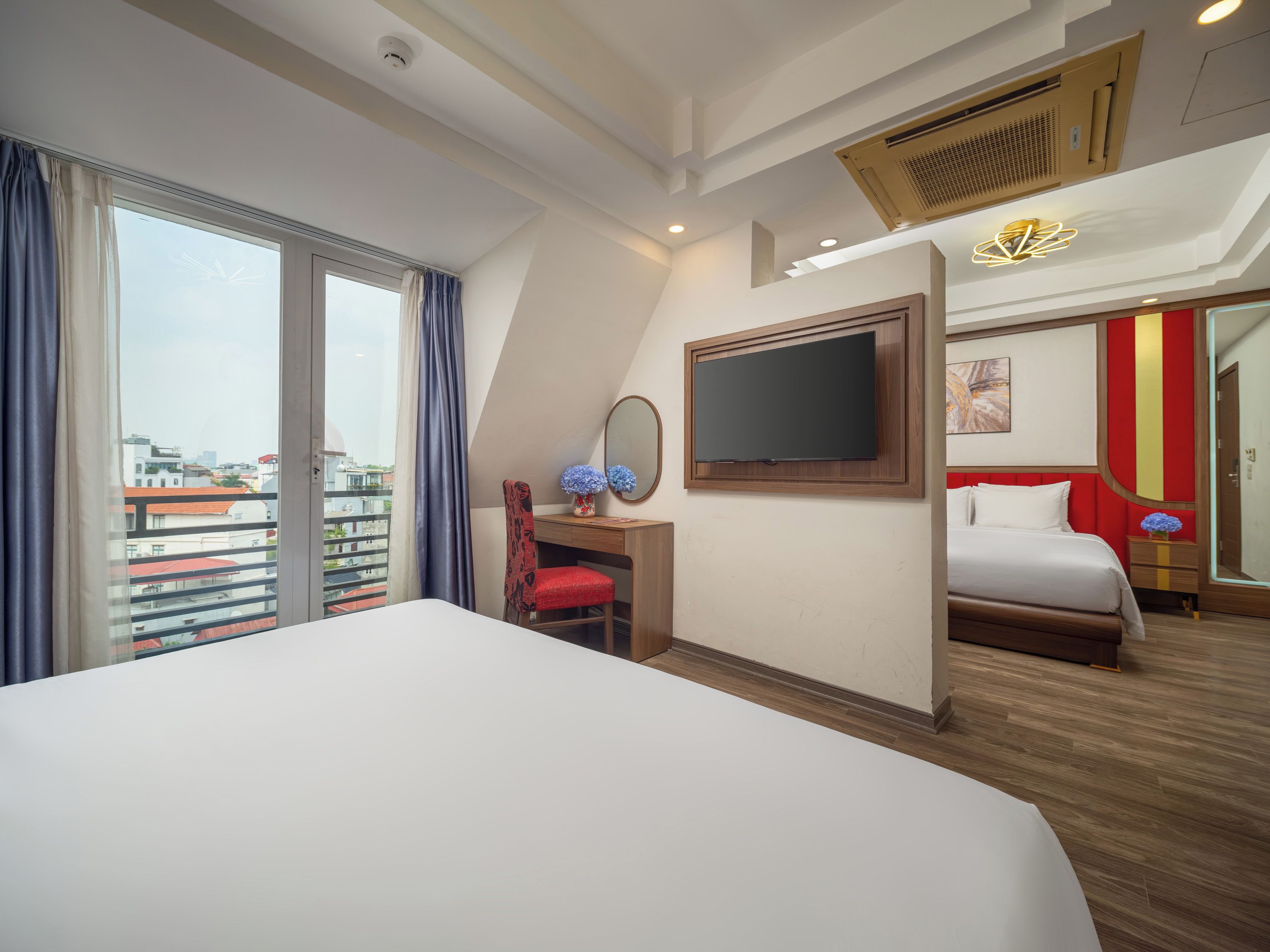 A hotel room with a large bed, a desk, a chair, a TV, and a balcony with a view of buildings.